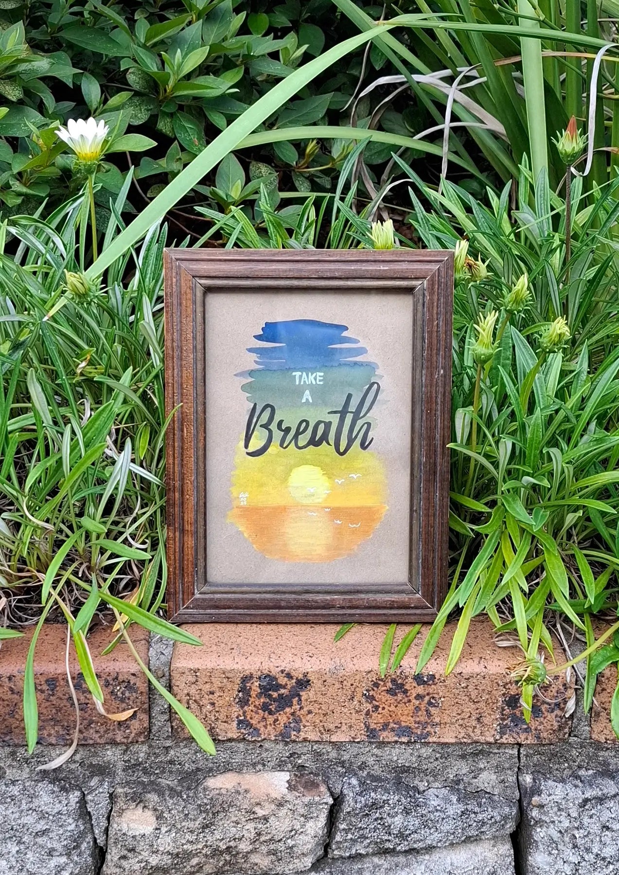 Framed artwork of serene gouache painting depicting a sunset on water on cardboard paper, with a background consisting of a gradient of blue to green, yellow and orange. Words on top say 'Take a Breath' in modern and cursive fonts. The small wooden frame sits on a brick wall with green plants in the background.