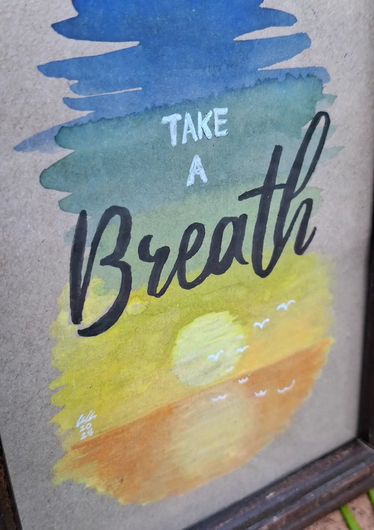Close up of artwork of serene gouache painting depicting a sunset on water on cardboard paper, with a background consisting of a gradient of blue to green, yellow and orange. Words on top say 'Take a Breath' in modern and cursive fonts with small white birds flying over the sunset.