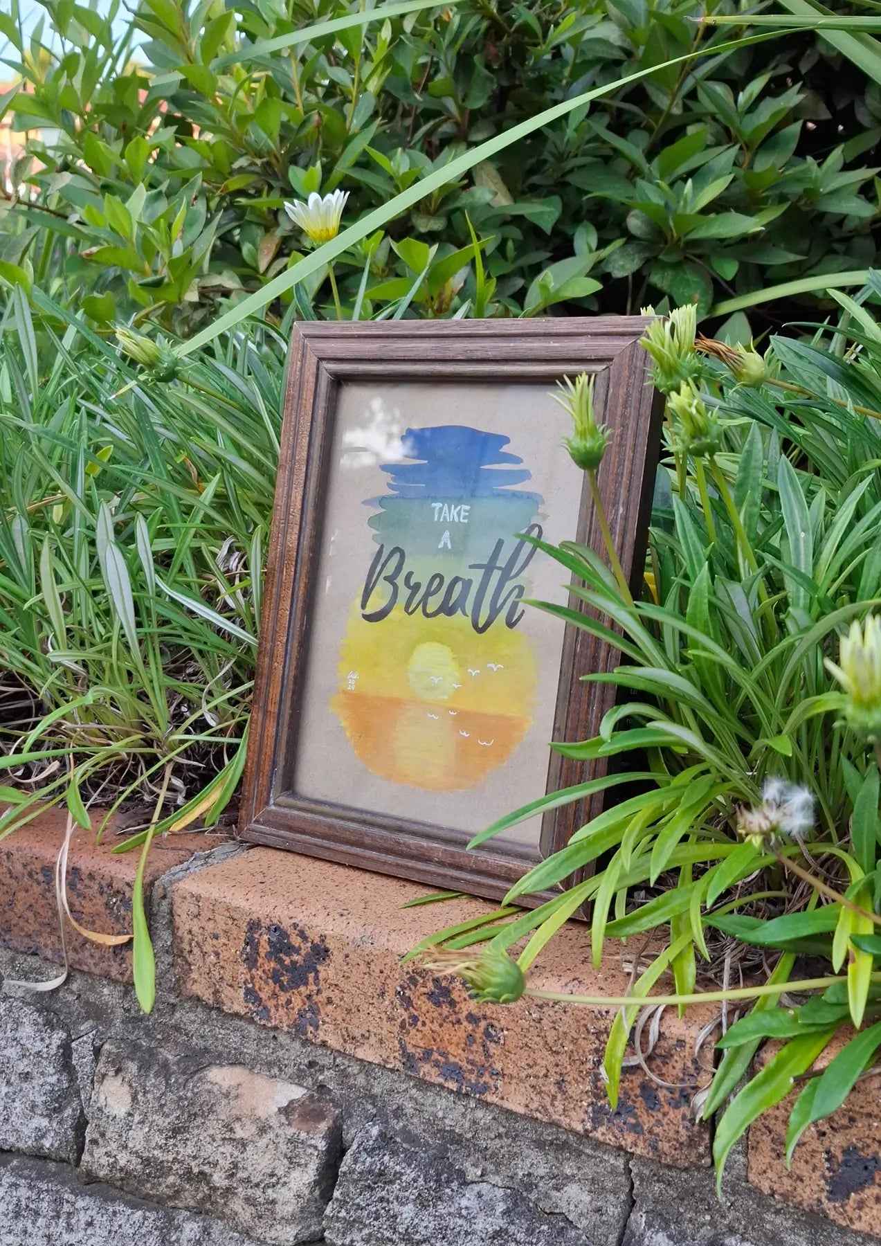 Framed artwork of serene gouache painting depicting a sunset on water on cardboard paper, with a background consisting of a gradient of blue to green, yellow and orange. Words on top say 'Take a Breath' in modern and cursive fonts. The small wooden frame sits on a brick wall with green plants in the background.