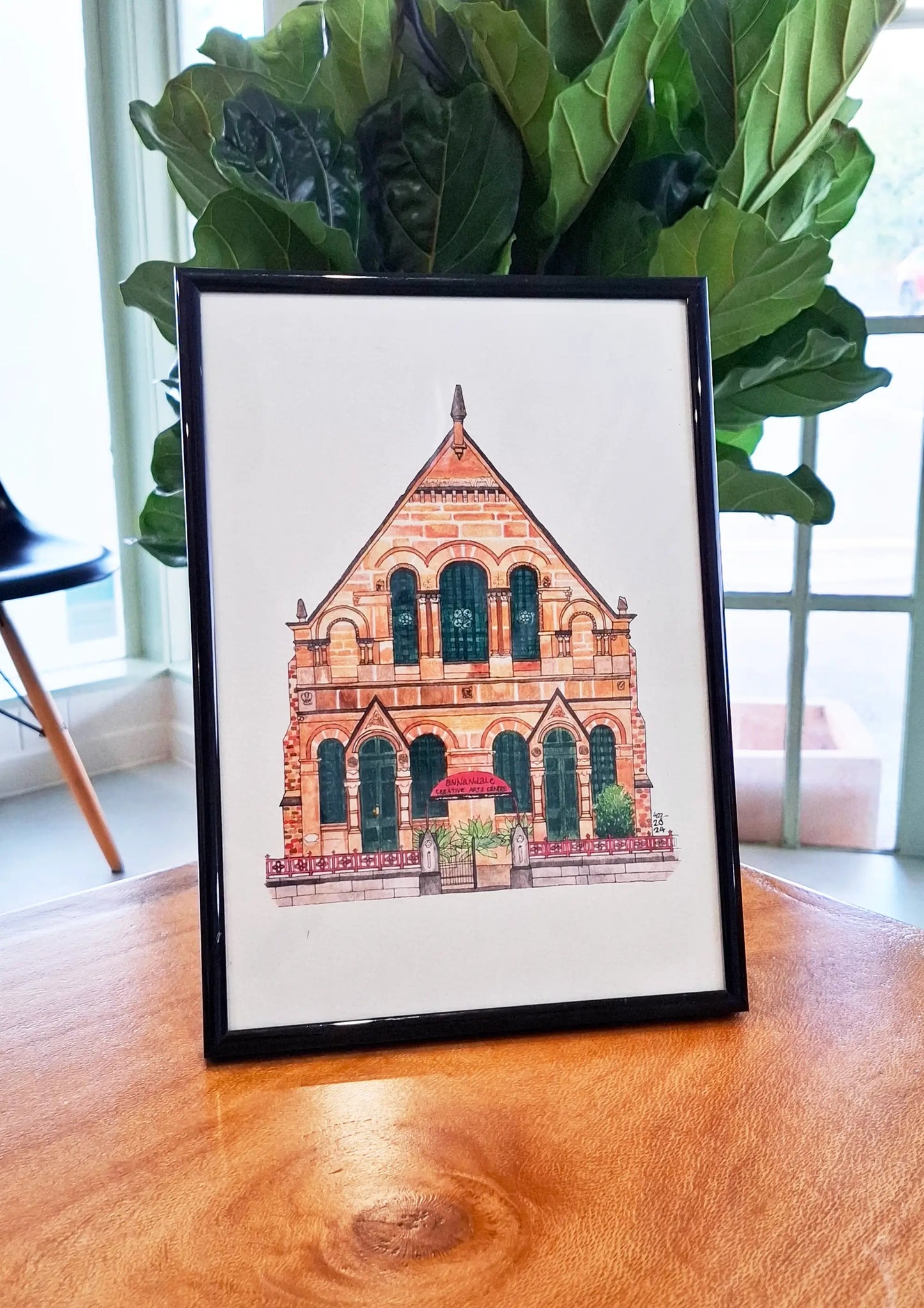 Print of ACAC illustration in a black frame sitting on a wooden table in a modern well lit room with fiddle leaf tree in background.