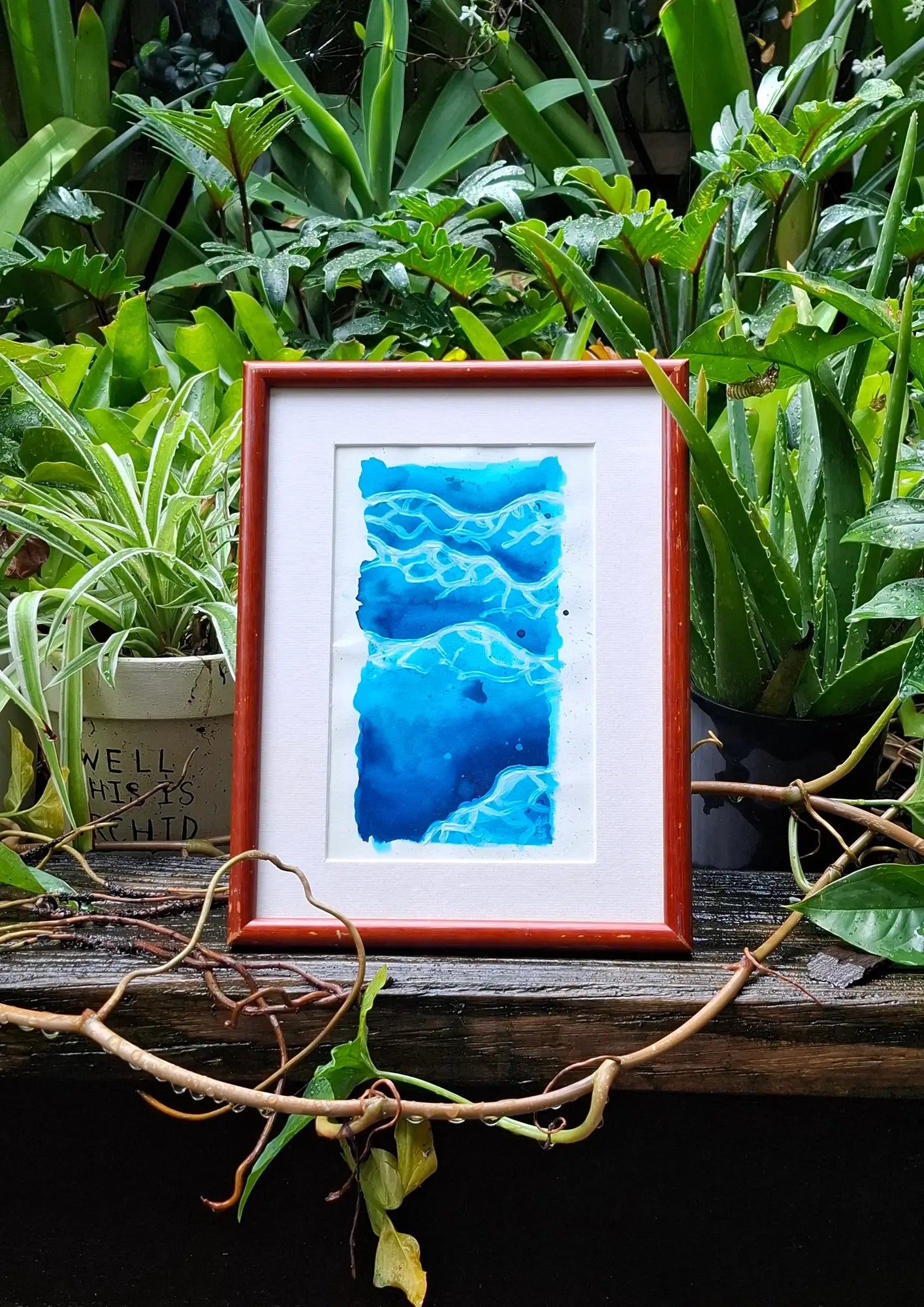 Framed artwork of serene gouache painting depicting gentle waves on textured paper, showcasing fluid movement and soft colours. Frame sits on wooden surrounded by green plants.