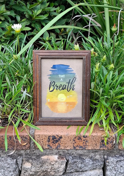 Framed artwork of serene gouache painting depicting a sunset on water on cardboard paper, with a background consisting of a gradient of blue to green, yellow and orange. Words on top say 'Take a Breath' in modern and cursive fonts. The small wooden frame sits on a brick wall with green plants in the background.