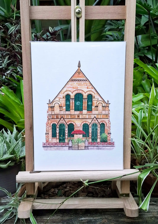 Print of ACAC illustration sitting on a small wooden easel in a garden with green leafy foliage in the background.